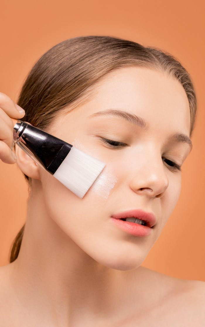 Close-up of a woman applying skincare product with a brush against a neutral background.