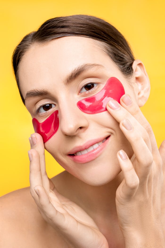 Woman smiling and applying red under eye masks for skincare routine.